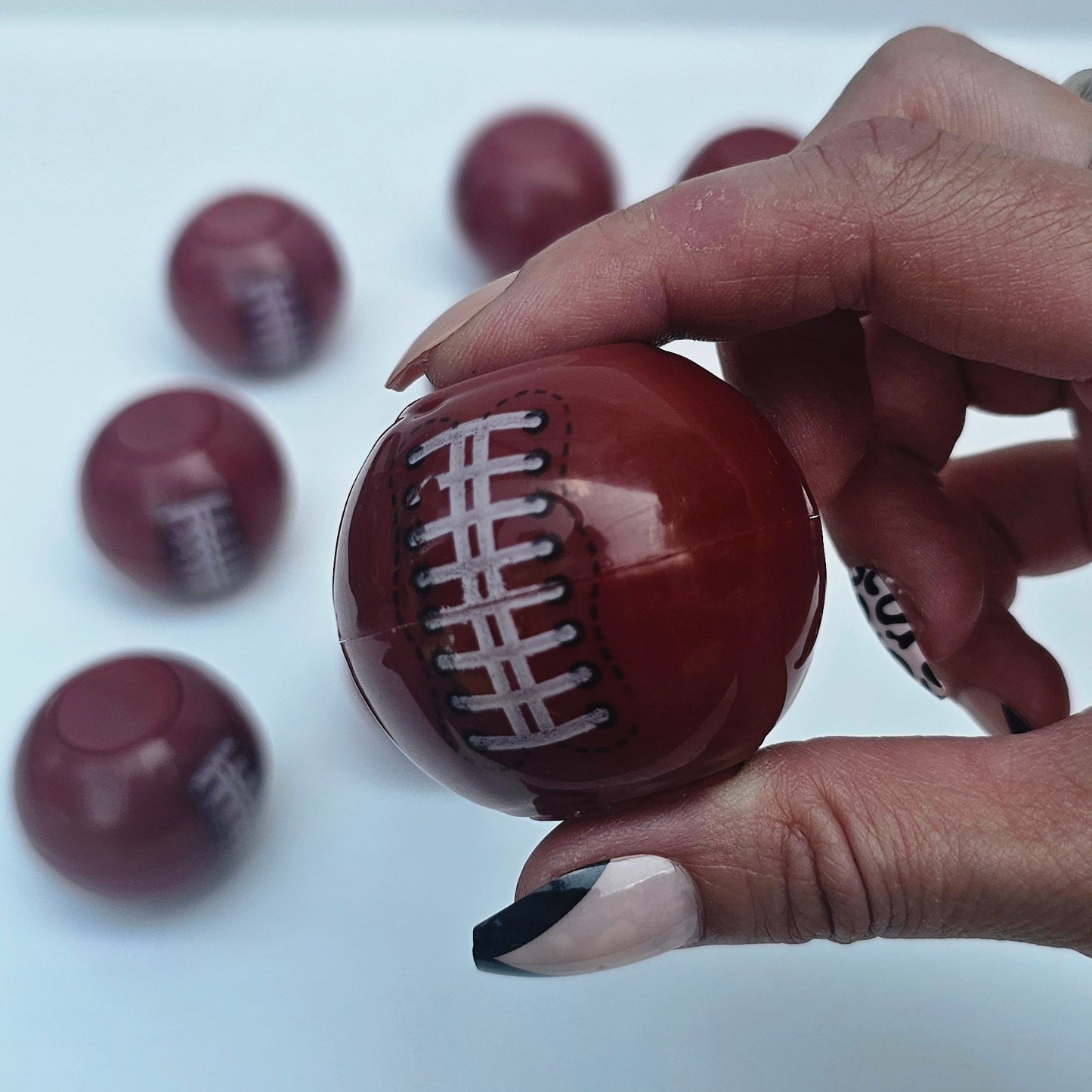 A hand with black and white painted fingernails holds a glossy, football-shaped brown candy or chocolate with white laces printed on it. Several identical football-shaped candies are scattered on a plain white surface in the background.