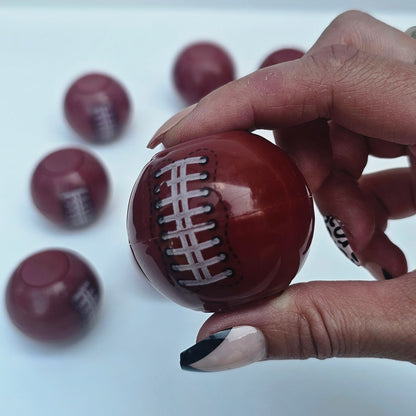 A hand with black and white painted fingernails holds a glossy, football-shaped brown candy or chocolate with white laces printed on it. Several identical football-shaped candies are scattered on a plain white surface in the background.